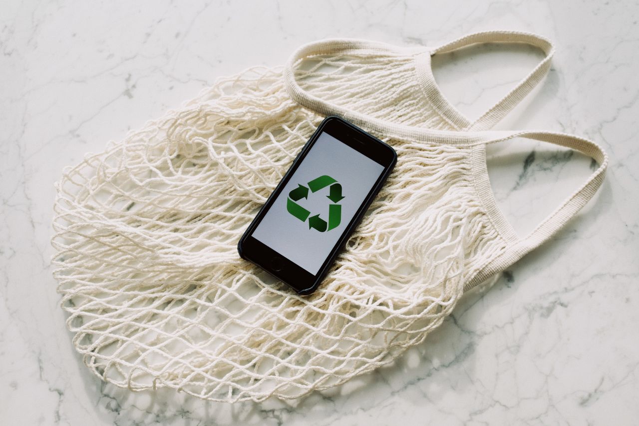 Overhead of smartphone with simple recycling sign on screen placed on white eco friendly mesh bag on marble table in room